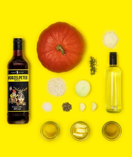 Ein roter Kürbis und eine Flasche Kräuterlikör stehen im Mittelpunkt, umgeben von Reis, Zwiebel und Gewürzen.