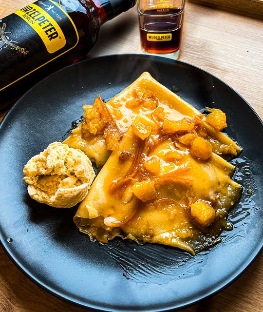 Leckere Pfannkuchen auf einem schwarzen Teller, überzogen mit einer süßen Sauce und frischen Früchten, begleitet von einem Likörglas.