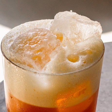 Das Getränk in einem klaren Glas zeigt eine lebendige orangefarbene Flüssigkeit, gekrönt von einer schaumigen Schicht.