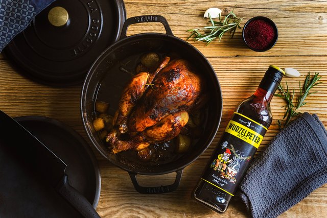 Neben dem Hähnchen steht eine Flasche Wurzelpeter, ein Kräuterlikör, und frische Rosmarinzweige liegen auf dem Holzbrett.