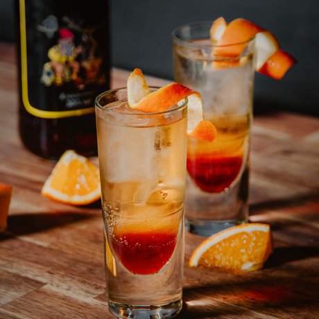 Auf einem rustikalen Tisch stehen zwei Cocktails, einer mit einer orangefarbenen Garnitur, umgeben von frischen Orangenstücken.