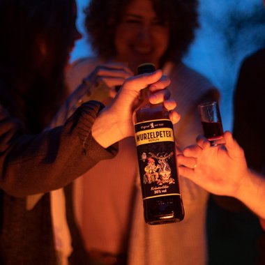 In der Dämmerung halten mehrere Personen eine Flasche Wurzelpeter und stoßen mit kleinen Gläsern an, um zu feiern.