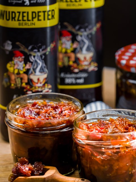 Zwei Gläser mit würzigem Aufstrich stehen auf einem Holztisch, umgeben von Flaschen mit dem Wurzelpeter-Label.