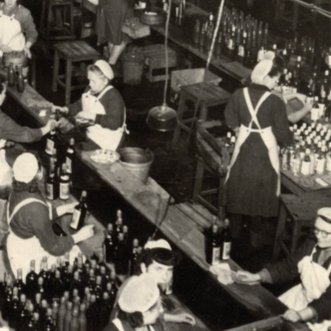 Die Szene zeigt eine Gruppe von Frauen, die in einer Produktionsstätte Flaschen abfüllen und sorgfältig etikettieren.