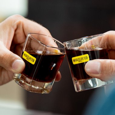 Zwei Hände stoßen mit Gläsern an, die mit Wurzelpeter beschriftet sind, während sie einen feierlichen Moment teilen.