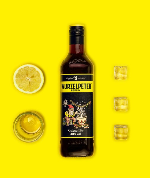 Die Flasche Wurzelpeter Kräuterlikör, eine Zitrone und Eiswürfel stehen auf einem knallgelben Hintergrund, der die Farben lebendig macht.