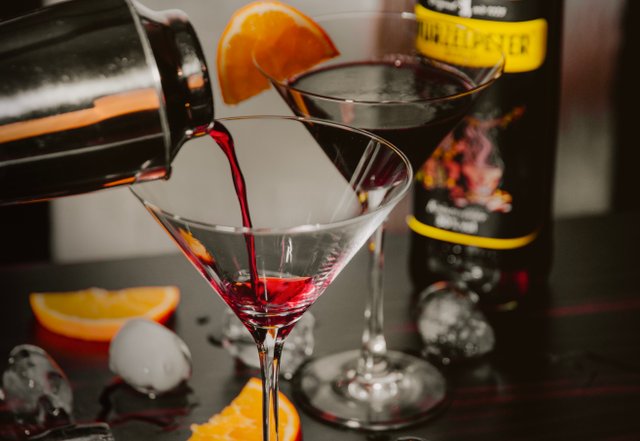 Ein Cocktailshaker gießt roten Likör in ein elegantes Martini-Glas, während ein zweites Glas daneben steht.