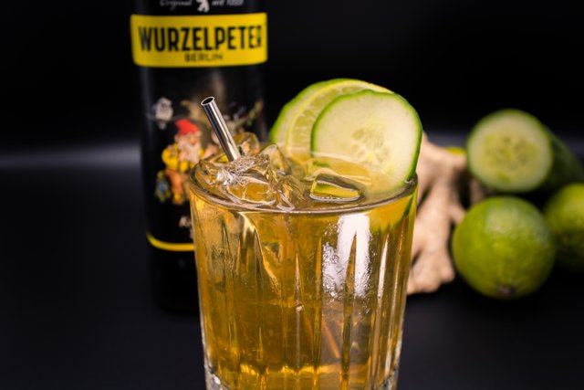 Ein Glas mit einem goldenen Getränk, umgeben von frischen Limetten und einer Flasche Wurzelpeter im Hintergrund.