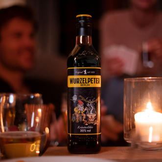Eine Flasche Wurzelpeter steht auf einem Tisch, umgeben von Gläsern und einer Kerze, während Menschen im Hintergrund lachen.