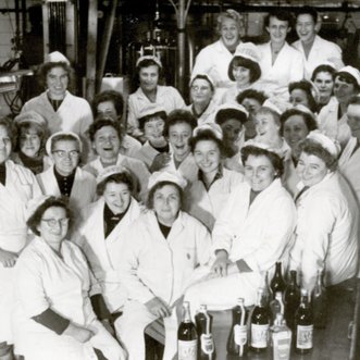 Eine Gruppe von Frauen in weißen Kitteln lächelt fröhlich in einer Fabrik, umgeben von Flaschen und Maschinen.