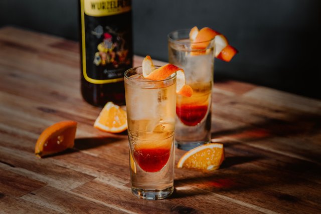 Auf einem rustikalen Tisch stehen zwei Cocktails, einer mit einer orangefarbenen Garnitur, umgeben von frischen Orangenstücken.