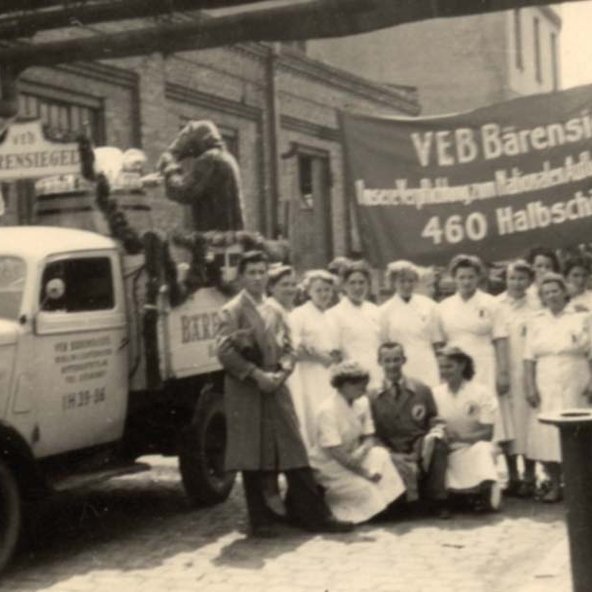 Die Szene zeigt eine Gruppe von Menschen in Arbeitskleidung, die vor einem dekorierten Lastwagen mit einem großen Schild stehen.