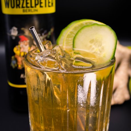 Ein Glas mit einem goldenen Getränk, umgeben von frischen Limetten und einer Flasche Wurzelpeter im Hintergrund.