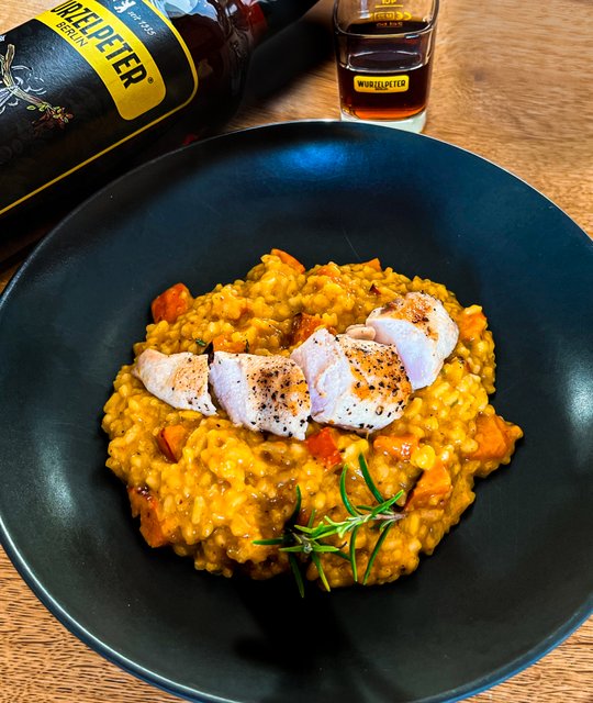 Das Risotto in der Schüssel ist leuchtend gelb, mit bunten Gemüsestücken und perfekt gebratenem Hähnchen, daneben ein Glas Wurzelpeter.
