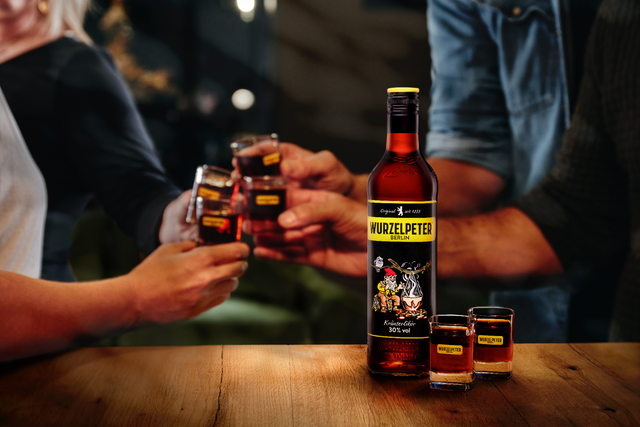 Eine Flasche Wurzelpeter Kräuterlikör steht auf einem Holztisch, umgeben von Gläsern, während Freunde anstoßen.