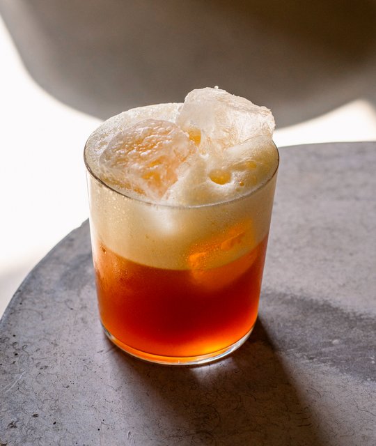 Das Getränk in einem klaren Glas zeigt eine lebendige orangefarbene Flüssigkeit, gekrönt von einer schaumigen Schicht.