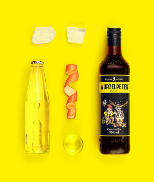 Die Szene zeigt eine Flasche Kräuterlikör, ein Glas, Eiswürfel und eine spiralförmige Orangenschale auf einem gelben Untergrund.